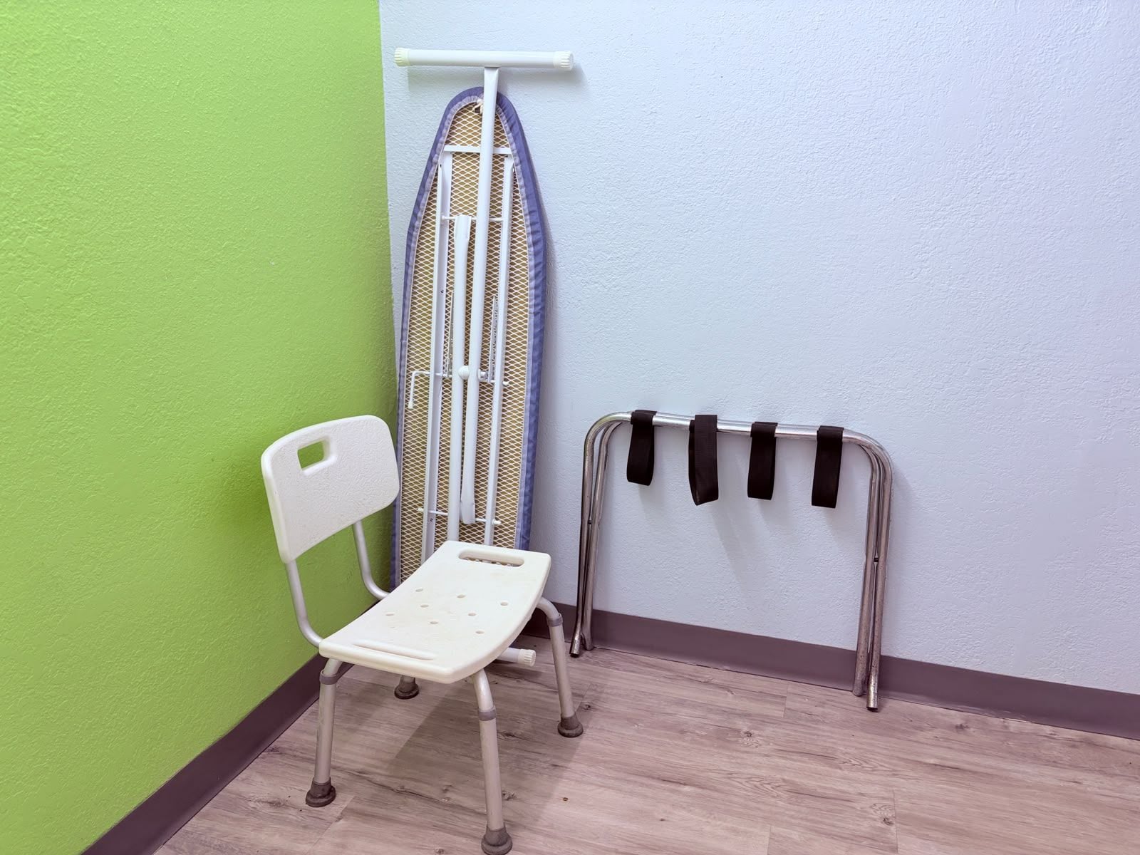 Eine Ecke eines Raumes mit einem weißen Klappstuhl, einem Metallbügelbrett und einem Metallgestell mit schwarzen Riemen.