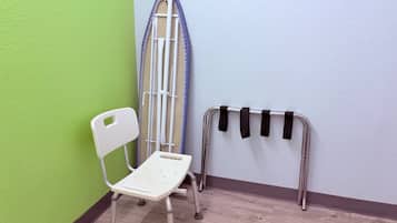 Eine Ecke eines Raumes mit einem weißen Klappstuhl, einem Metallbügelbrett und einem Metallgestell mit schwarzen Riemen.