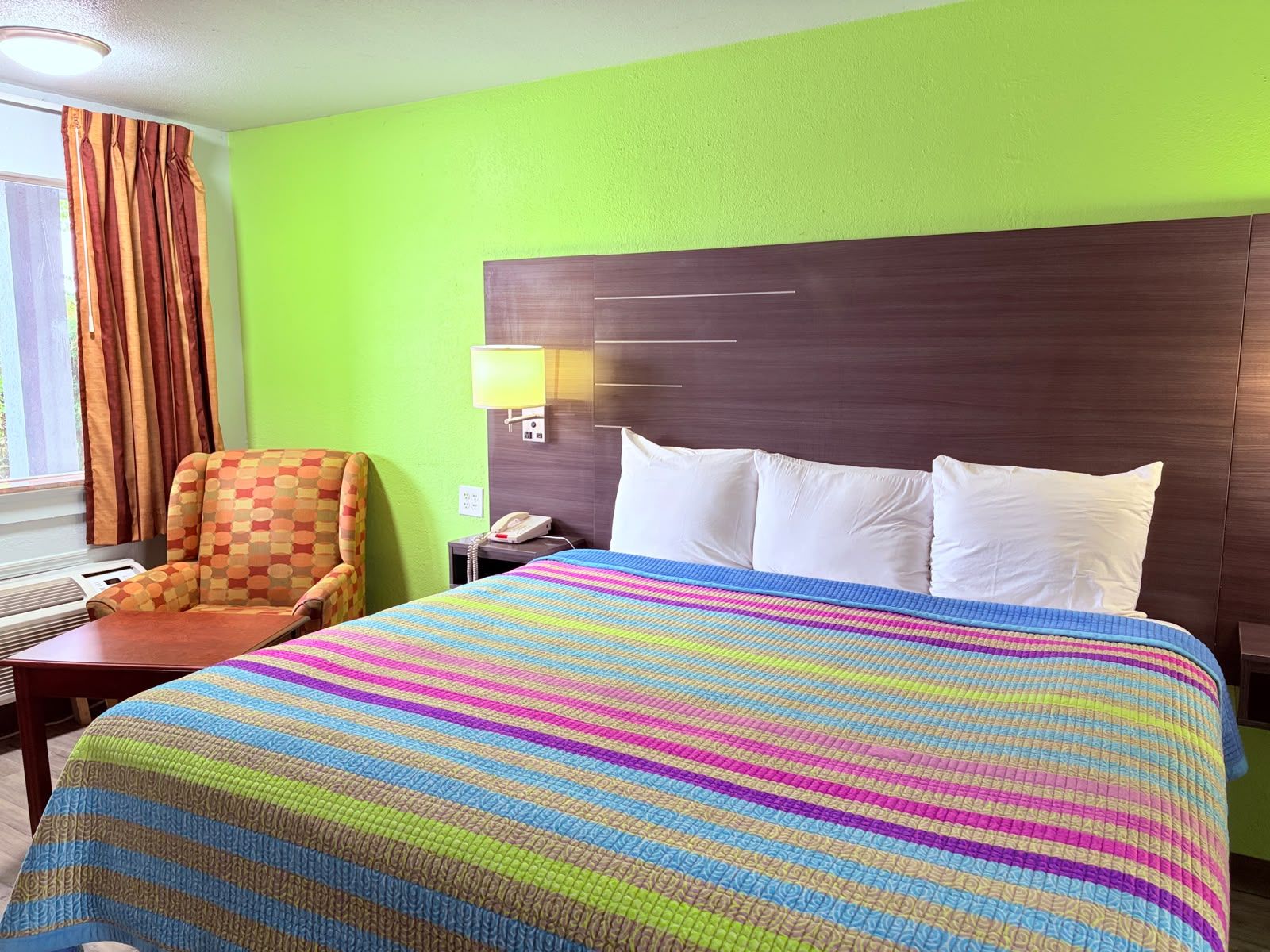 A hotel room with a bed, a chair, a nightstand, a lamp, and a window with curtains.