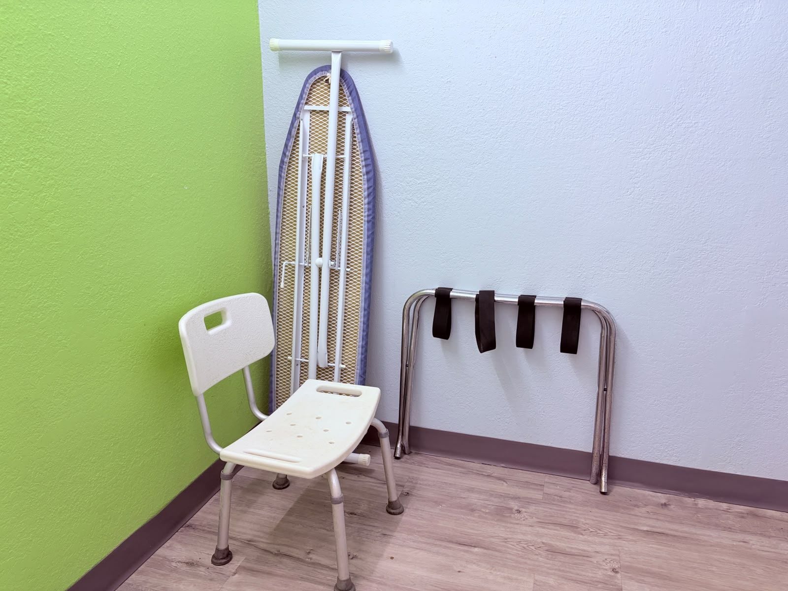 A corner of a room with a white chair, a metal ironing board, and a metal rack with black straps.