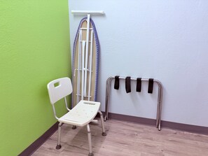 A corner of a room with a white chair, a metal ironing board, and a metal rack with black straps.