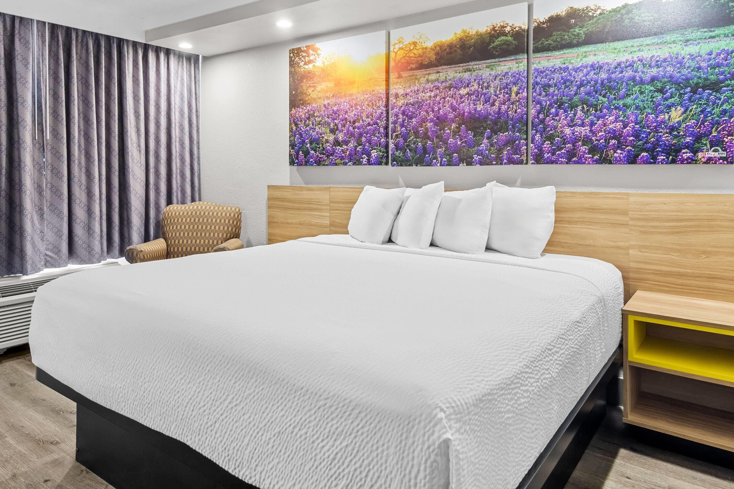A modern bedroom with a large bed, a headboard, a bedside table, a chair, and a wall with a floral painting.