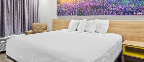 A modern bedroom with a large bed, a headboard, a bedside table, a chair, and a wall with a floral painting.