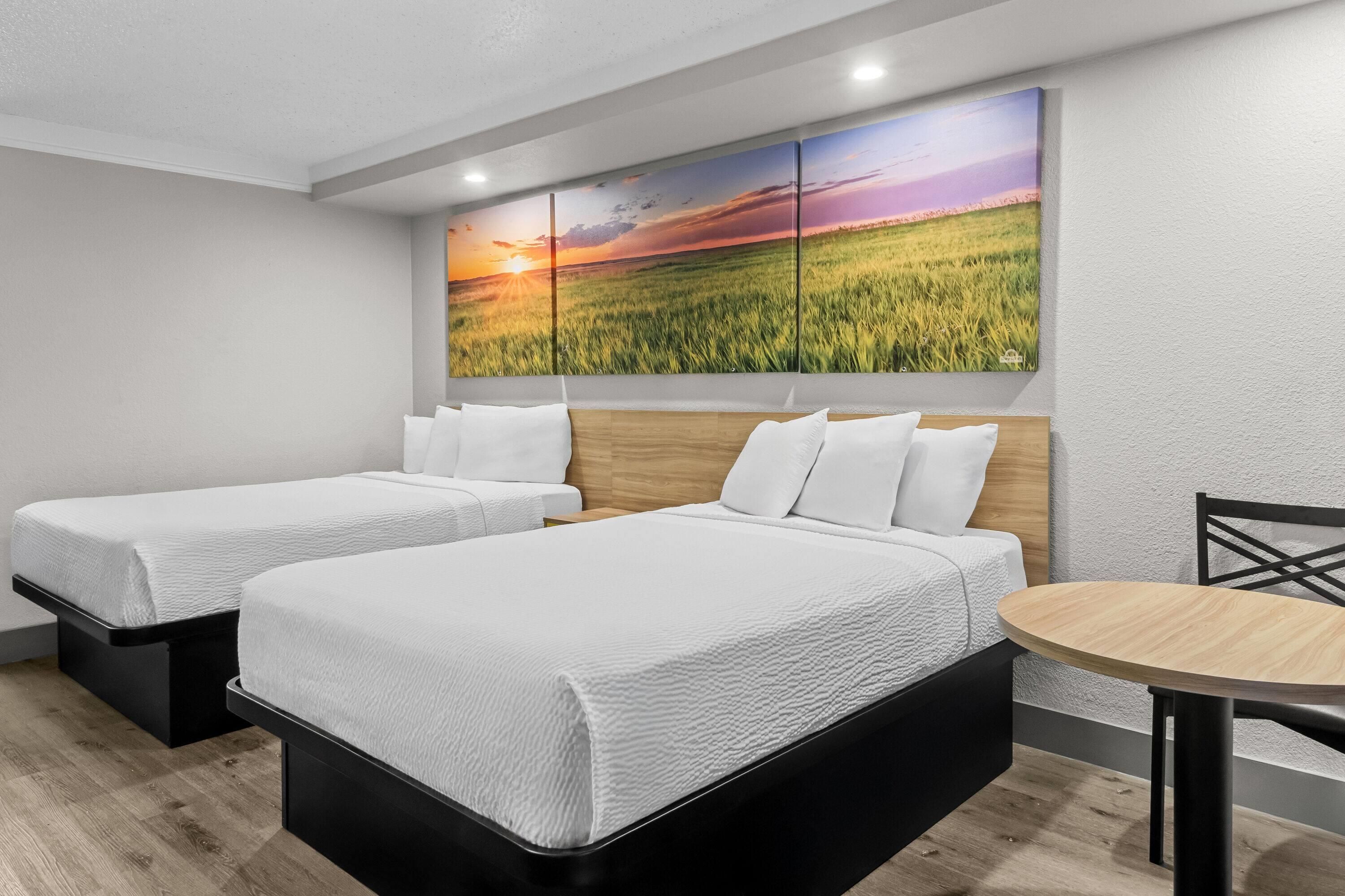 A hotel room with two beds, a wooden headboard, a round table, and a large landscape painting on the wall.