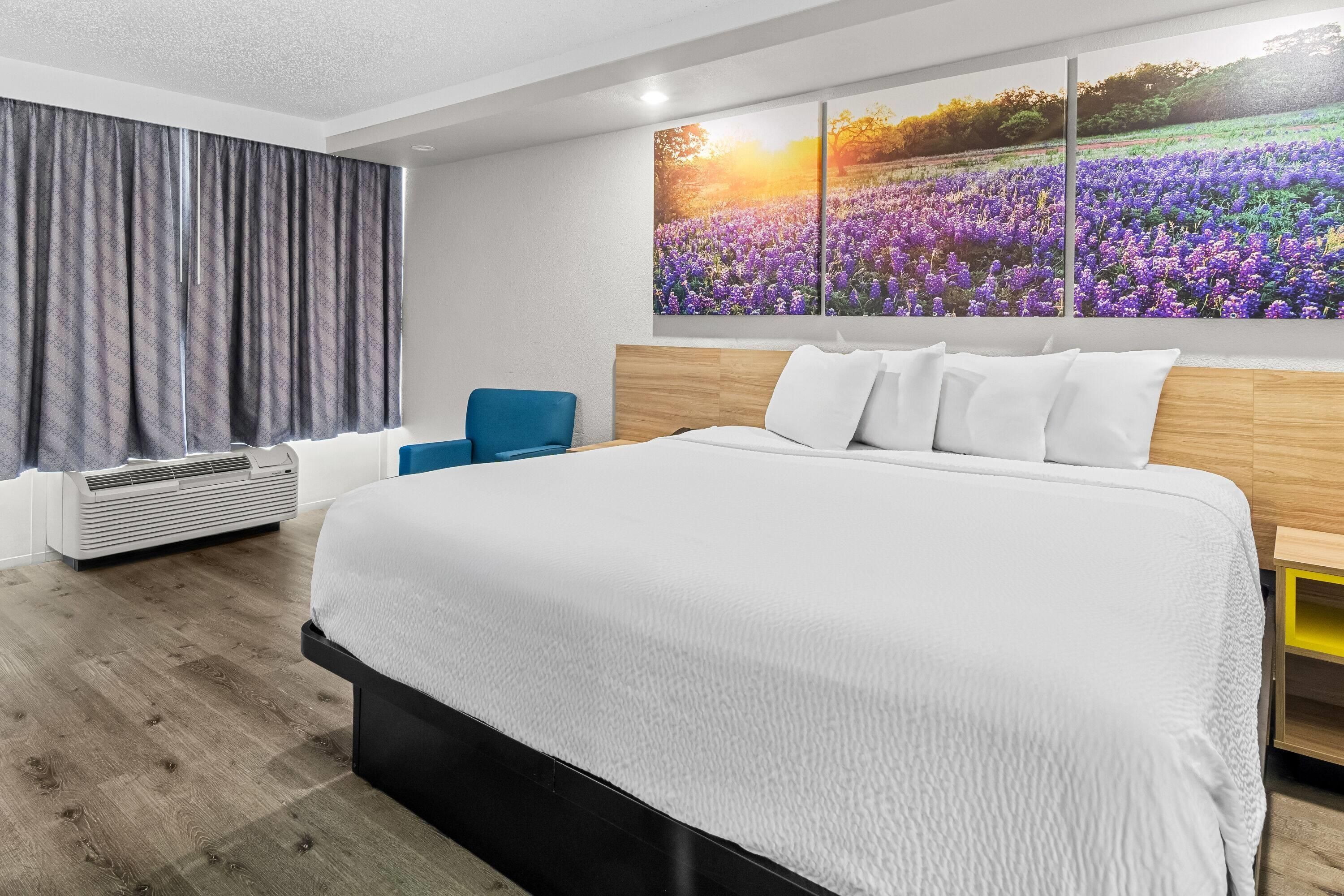 Une chambre moderne avec un grand lit, un fauteuil bleu, un climatiseur fixé au mur et une grande œuvre d’art à motifs floraux.