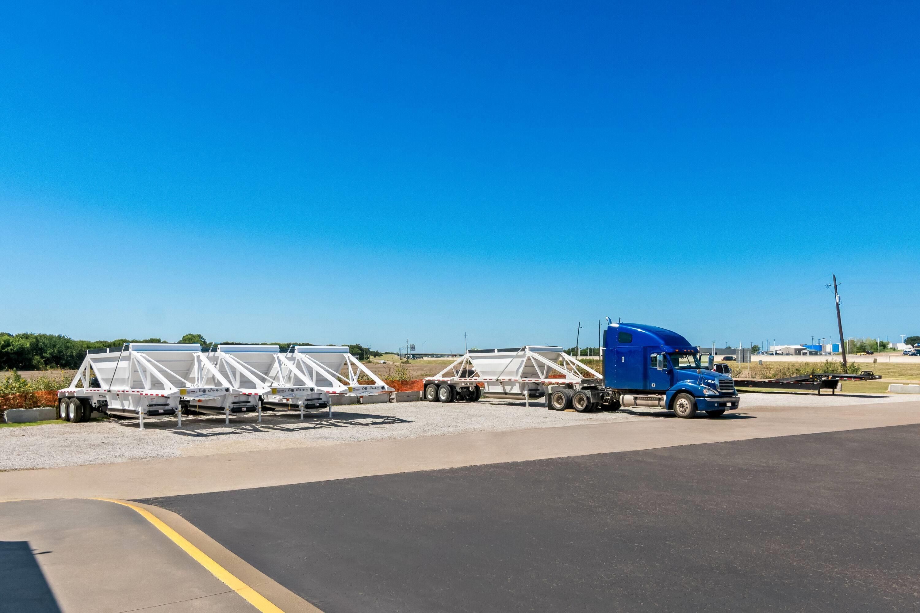 Un camión azul remolqueado estacionado en una carretera, transportando estructuras blancas, posiblemente paneles solares, bajo un cielo despejado.