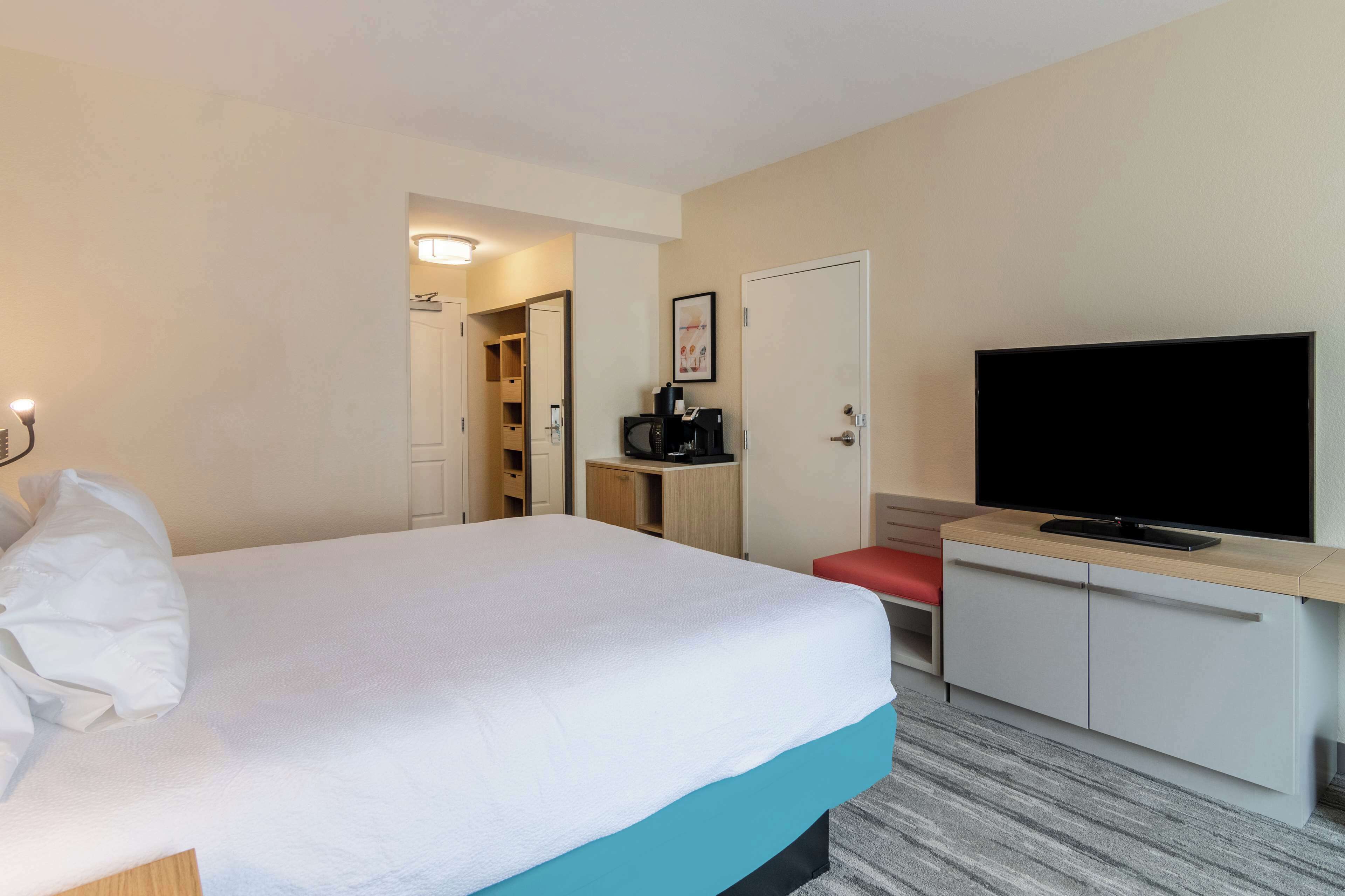 Habitación de hotel con cama, televisión, escritorio y cafetera, y puerta que da a un baño.