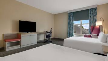 Ein Hotelzimmer mit zwei Betten, einem Schreibtisch mit Fernseher, einem Stuhl und einem Fenster mit Vorhängen.