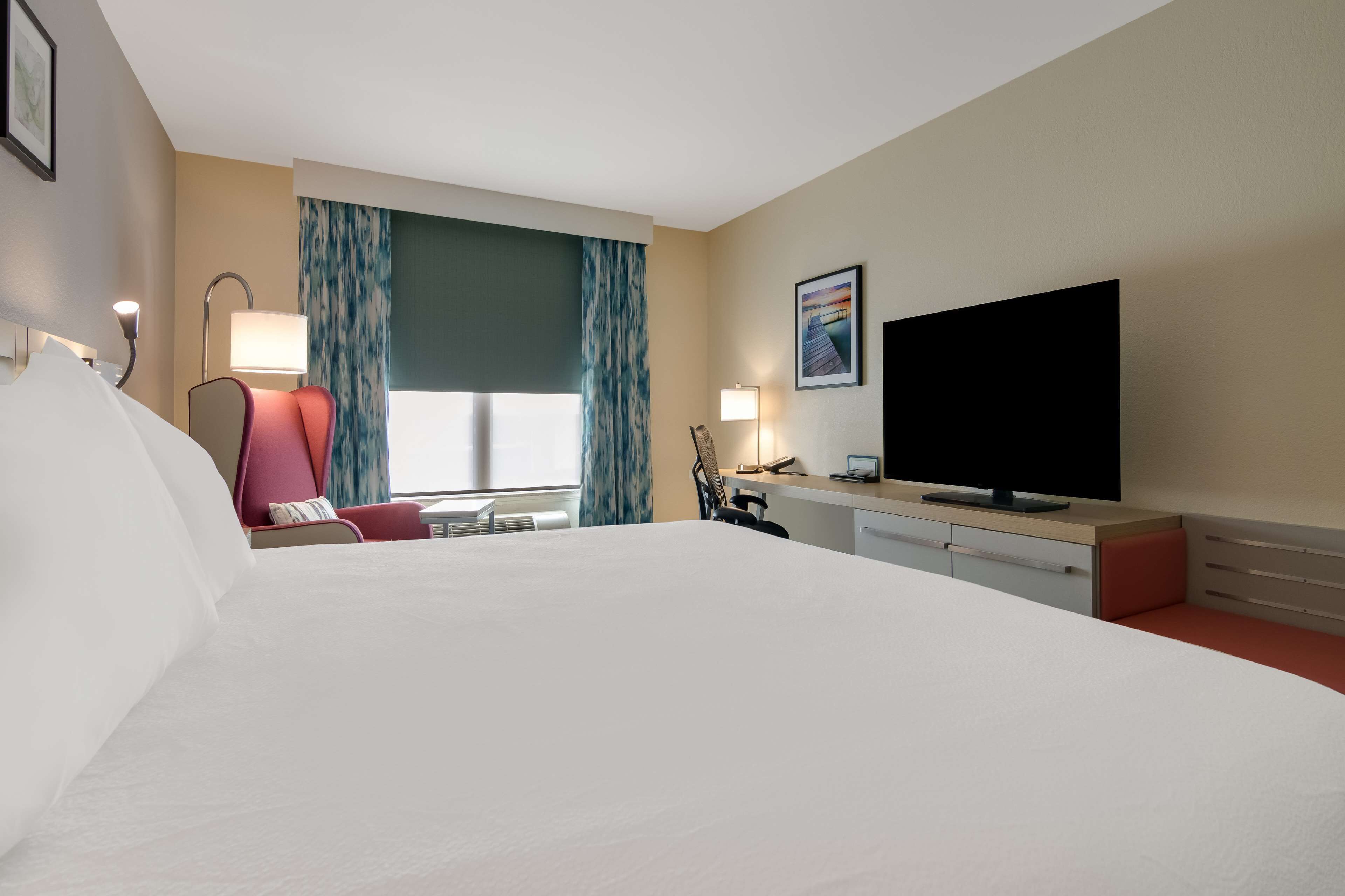Habitación de hotel con una cama grande, un escritorio, un televisor de pantalla plana, una silla y un ventanal con cortinas.