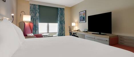Habitación de hotel con una cama grande, un escritorio, un televisor de pantalla plana, una silla y un ventanal con cortinas.