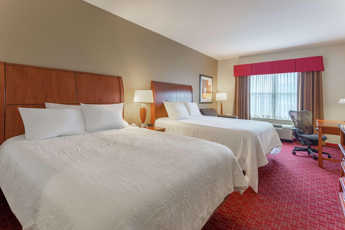 A hotel room with two beds, a desk, a chair, and a window with curtains.