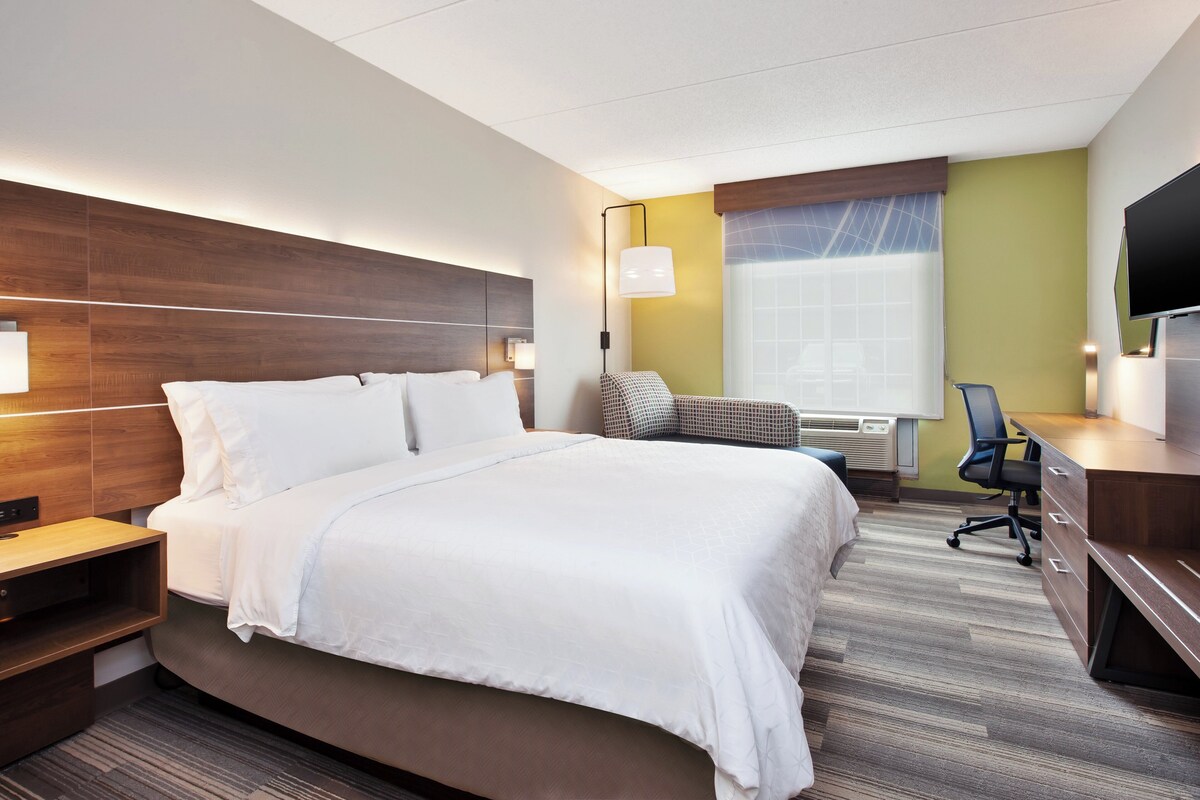 A hotel room with a large bed, a desk, a chair, a TV, and a window with blinds.