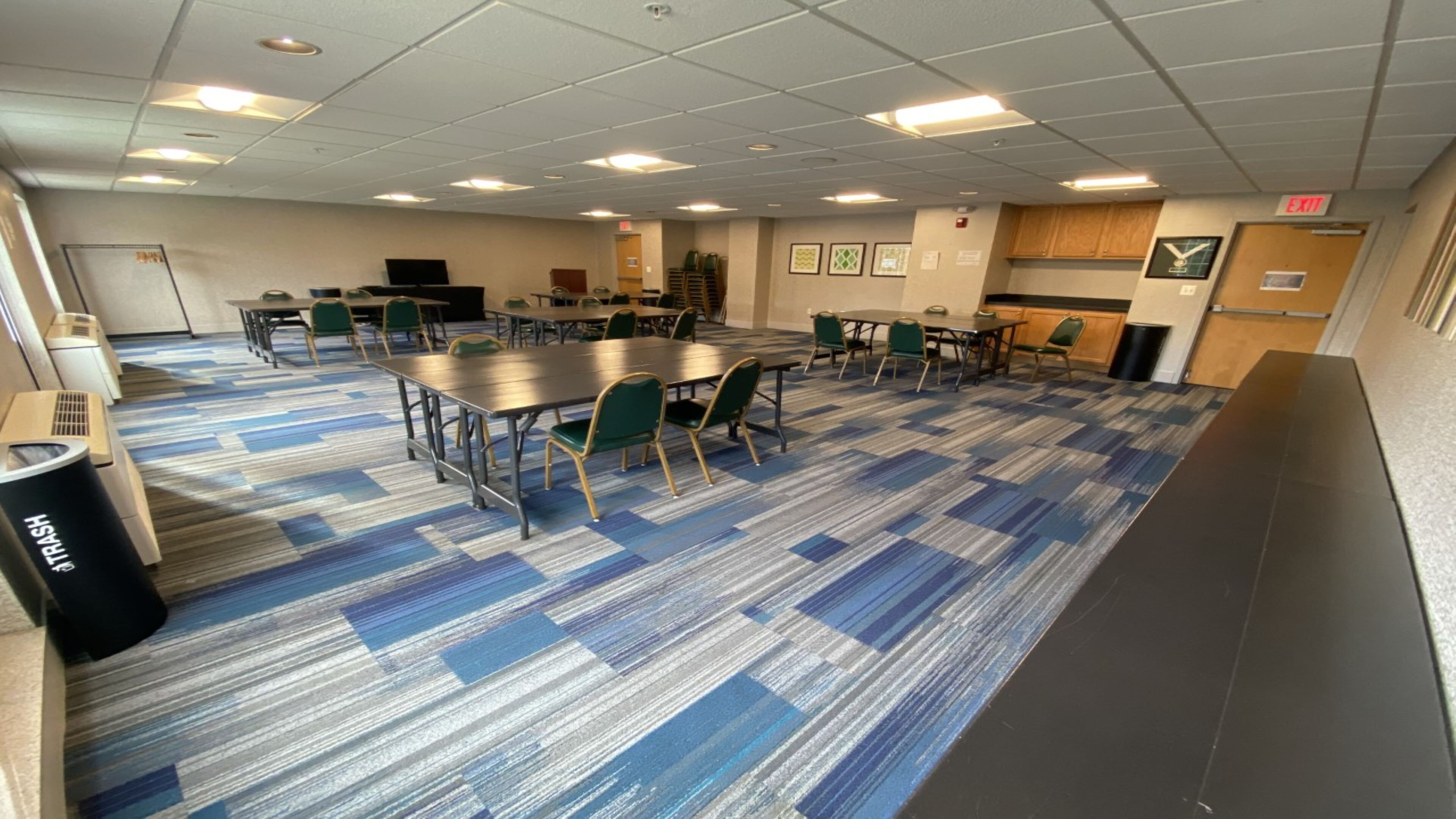 A conference room with tables and chairs, a trash can, and a kitchenette area.
