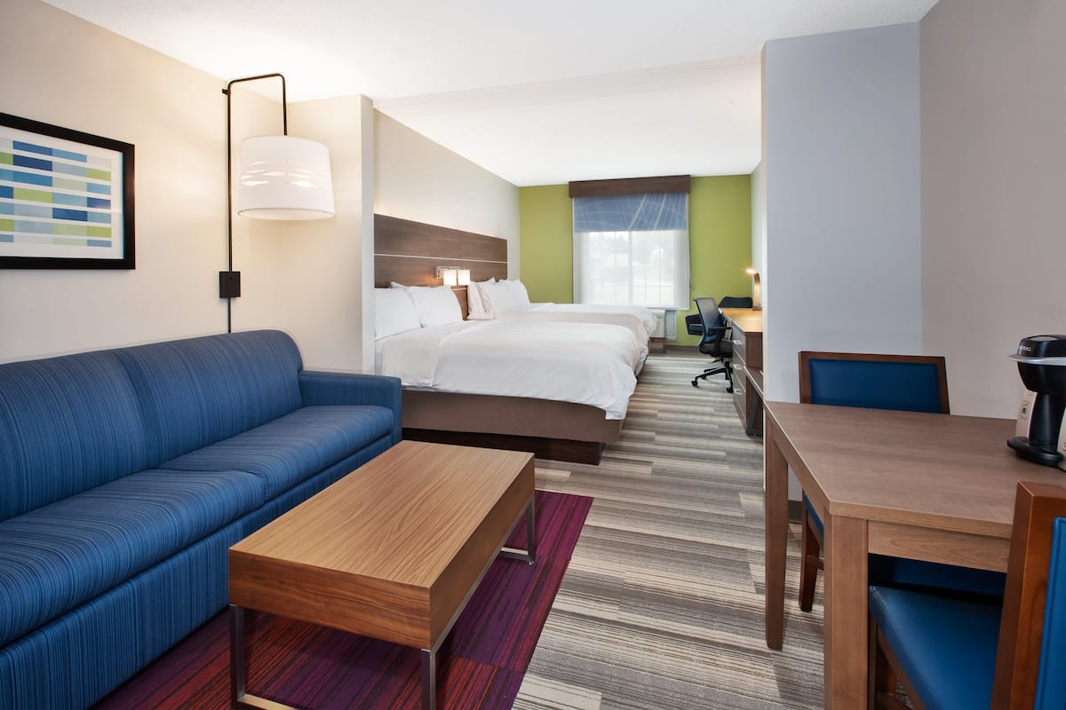 A hotel room with a blue sofa, a wooden coffee table, a bed with white linens, a desk with a chair, and a lamp.