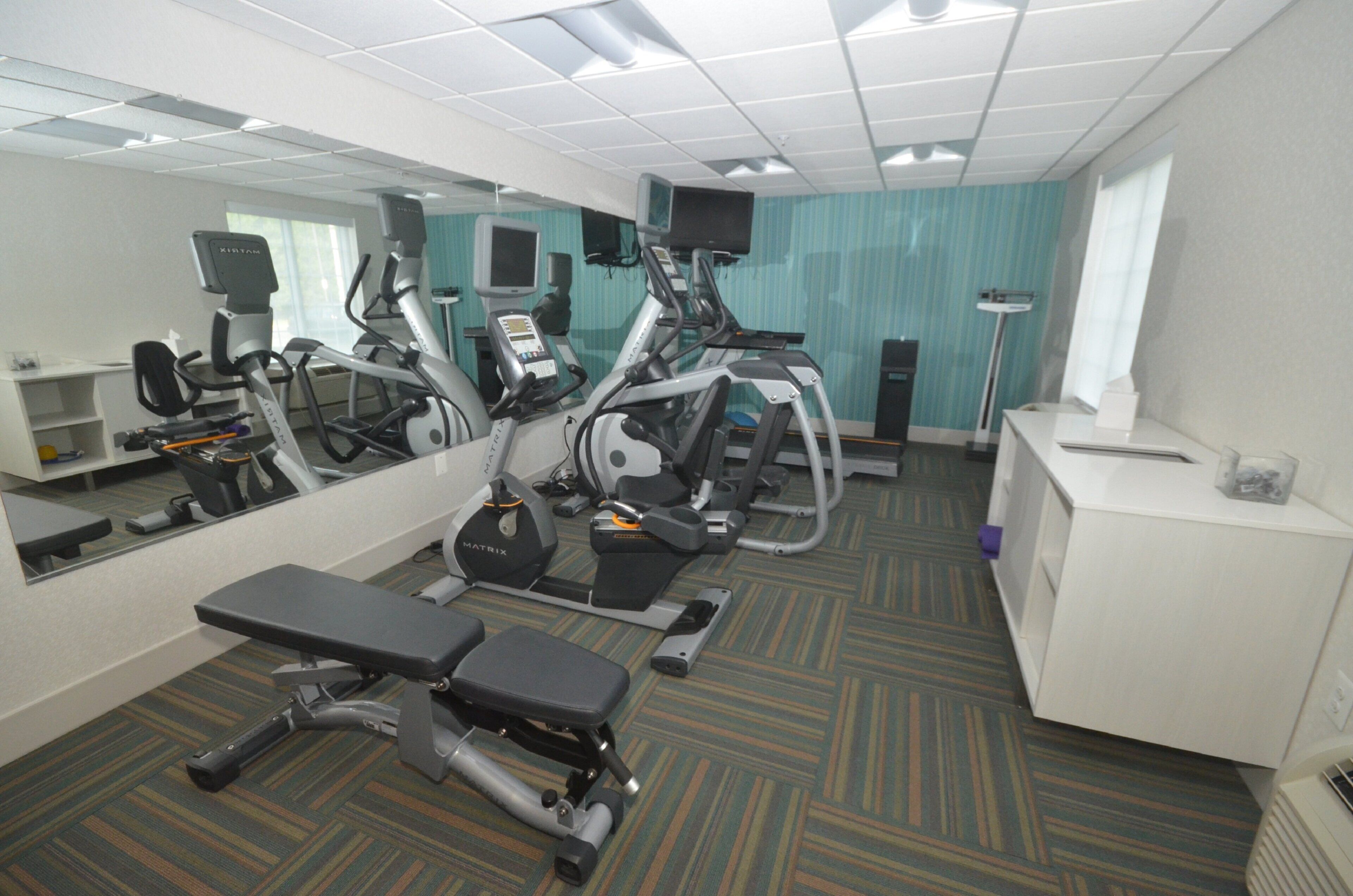 Sala de ginástica com equipamentos de exercício, espelho, escrivaninha e televisão.