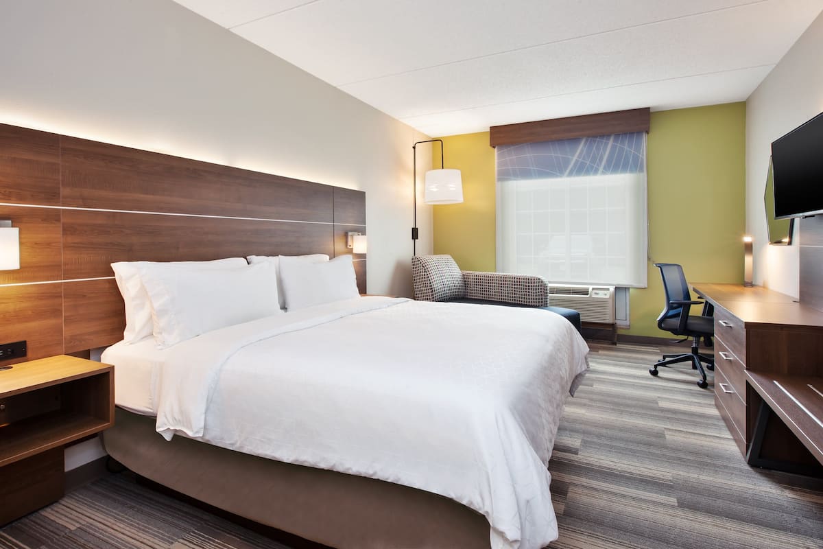 A hotel room with a large bed, a desk, a chair, a TV, and a window with blinds.