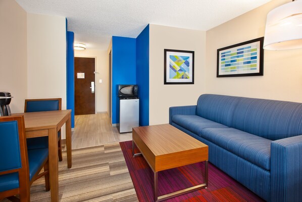 A modern hotel room with a blue sofa, wooden coffee table, and a dining area with a table and chairs. - Holiday Inn Express Hotel & Suites Warrenton by IHG (Warrenton)