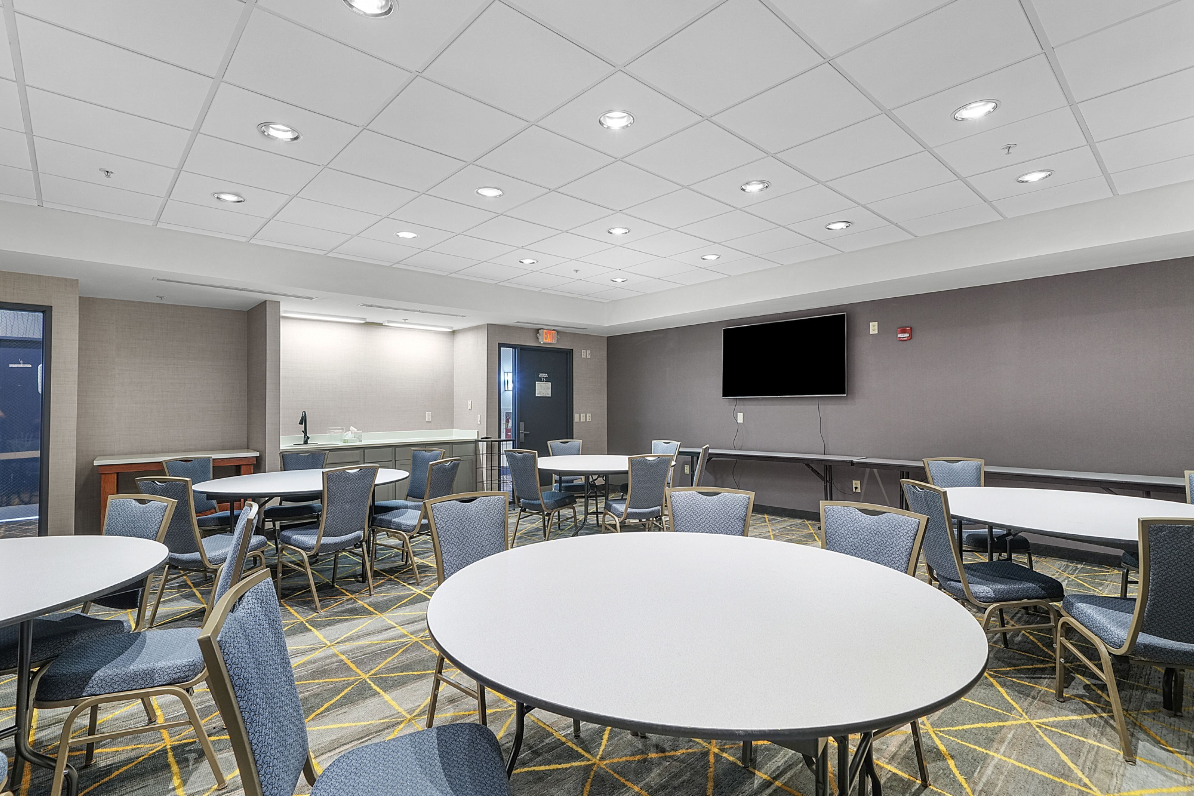 A conference room with multiple tables and chairs, a flat-screen TV mounted on the wall, and a door marked 'EXIT'.