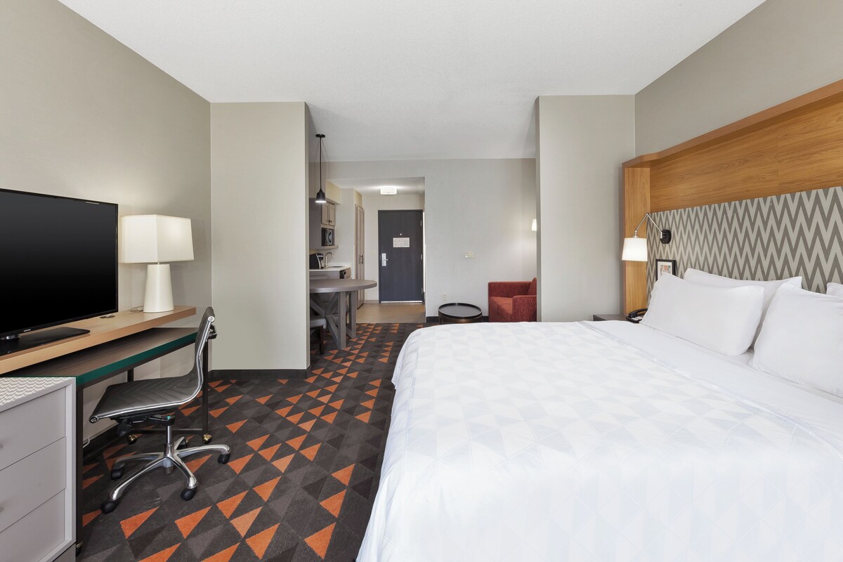 A hotel room with a bed, desk, chair, television, and a view of the hallway.