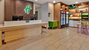 Una zona de recepción con un mostrador de madera, dos computadoras, un mueble verde para guardar objetos, un refrigerador con bebidas y una barra de aperitivos con asientos.