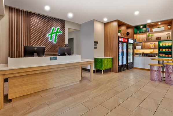 A reception area with a wooden reception desk, two computers, a green storage unit, a refrigerator with beverages, and a snack bar with seating. - Holiday Inn Grand Rapids - South by IHG (Grand Rapids)