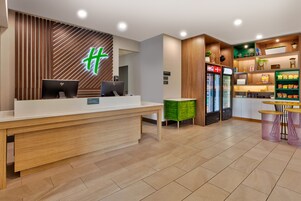 Una zona de recepción con un mostrador de madera, dos computadoras, un mueble verde para guardar objetos, un refrigerador con bebidas y una barra de aperitivos con asientos.