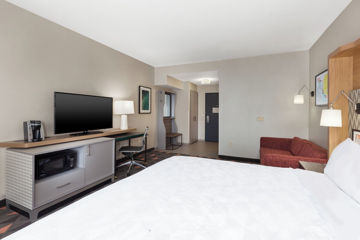 A hotel room with a bed, a TV, a microwave, a desk with a lamp, and a red chair.