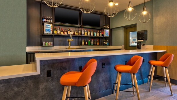 Una zona de bar con mostrador, taburetes y estantes llenos de botellas.