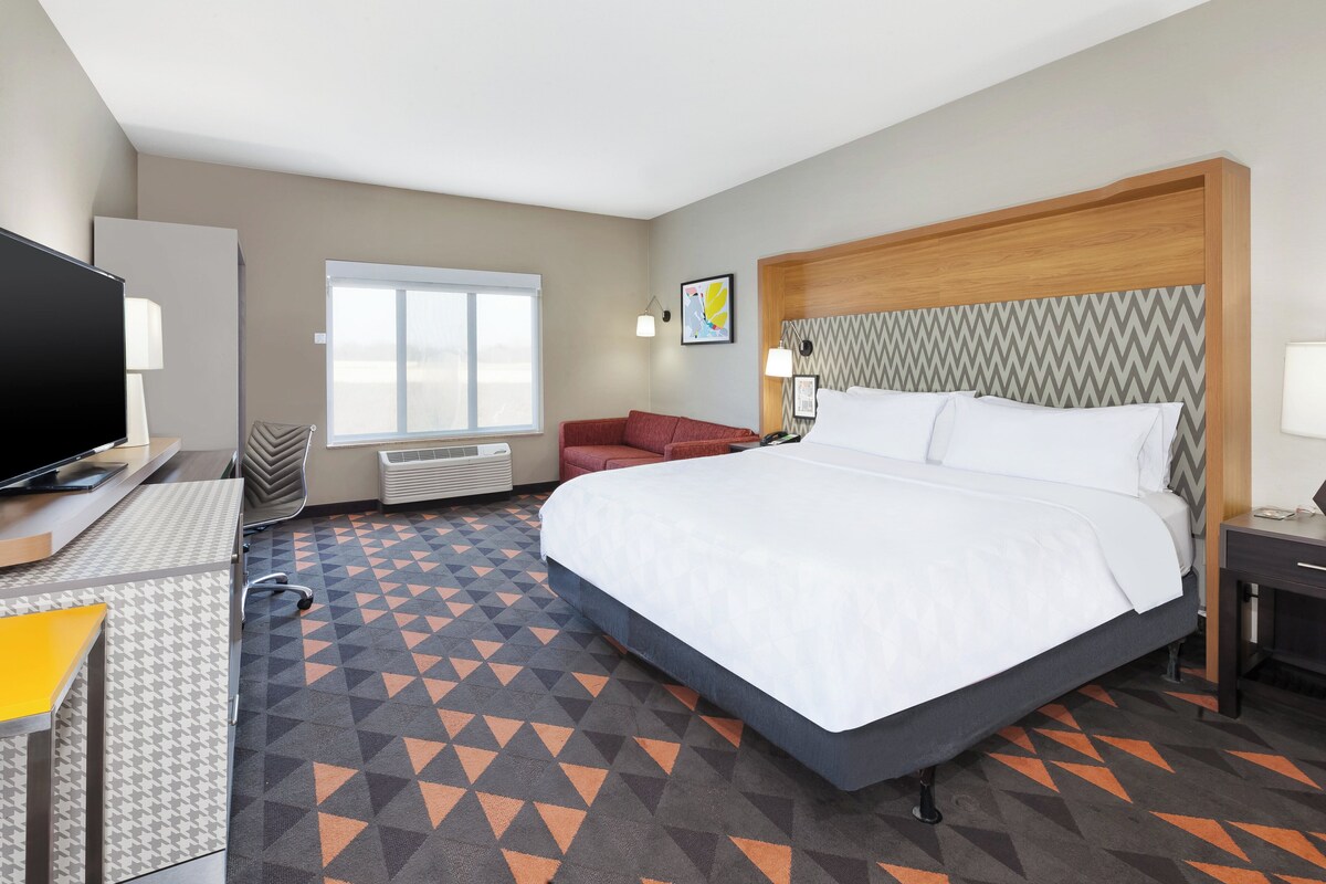 A hotel room with a large bed, a desk with a flat-screen TV, a chair, a sofa, and a window with a view.