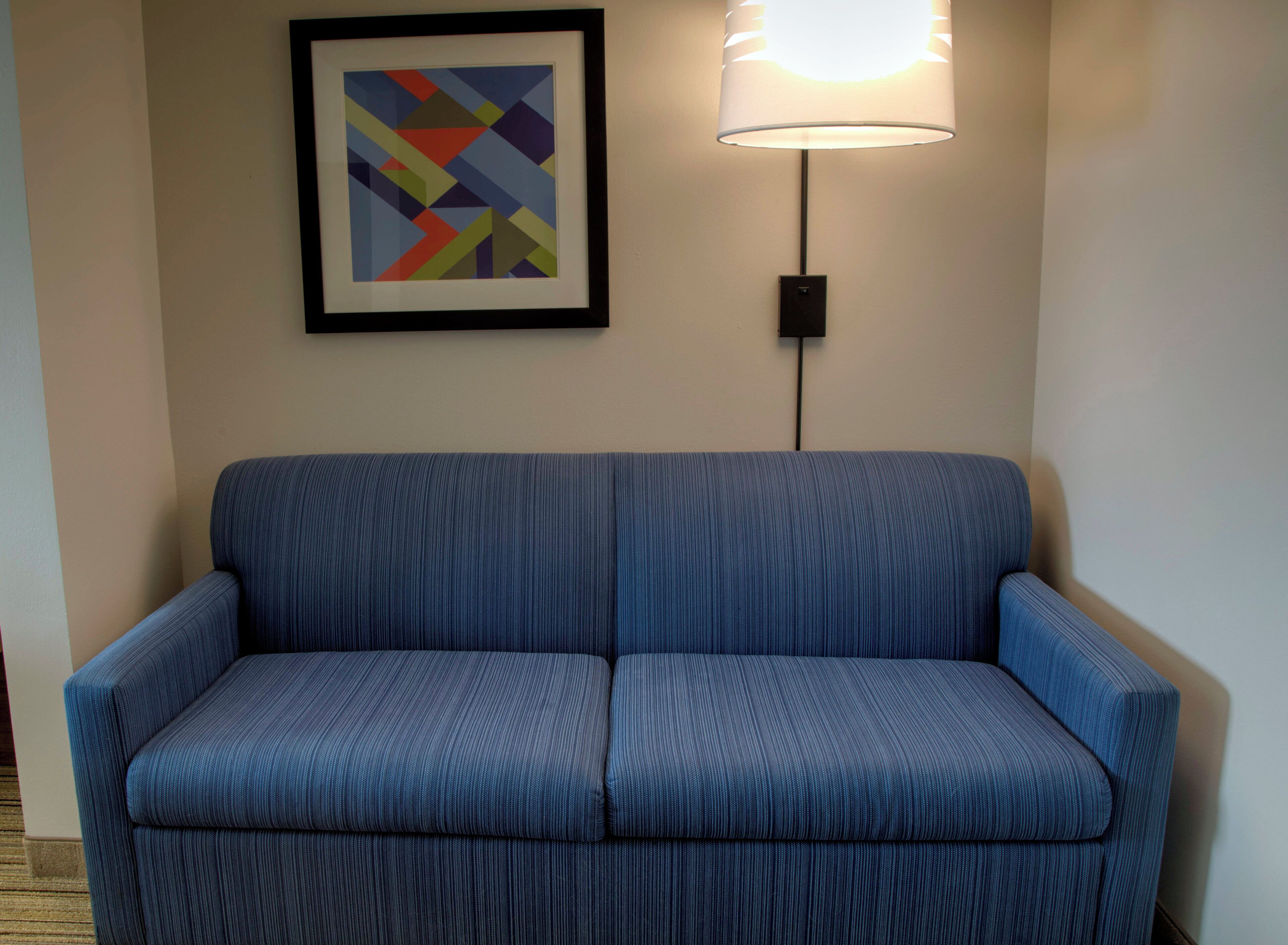 A blue sofa with a framed abstract painting above it and a wall-mounted lamp to the right.