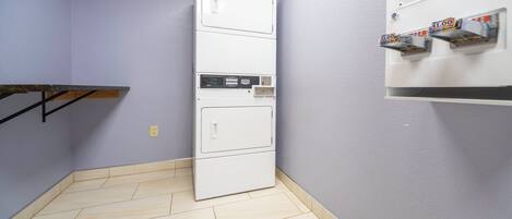 Una habitación con una máquina tragamonedas blanca fijada a la pared, un electrodoméstico blanco con pantalla digital y un estante de madera.