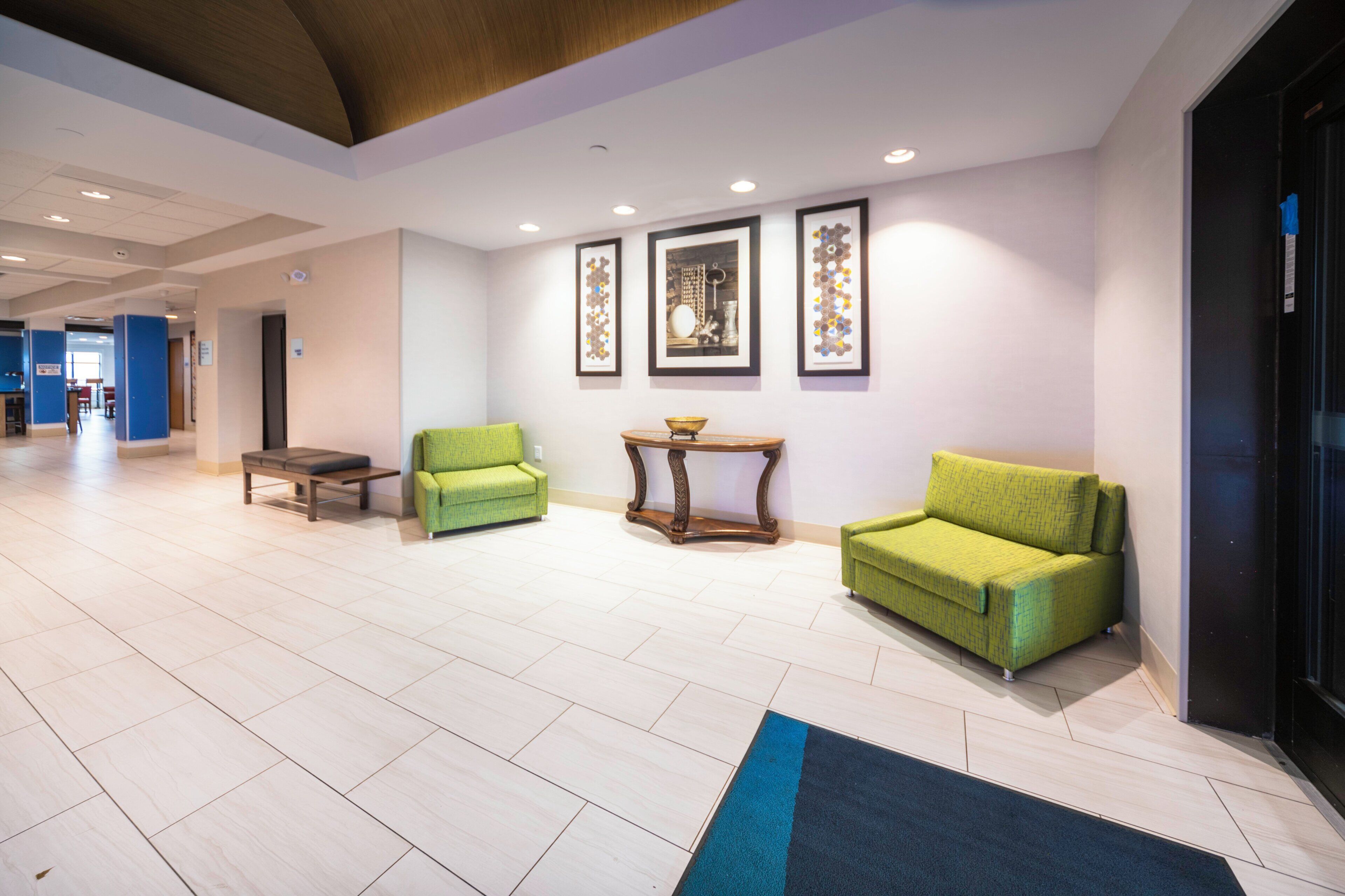 Un hall d’hôtel avec un comptoir d’accueil, deux fauteuils verts, une petite table sur laquelle se trouve un bol, et des œuvres d’art encadrées accrochées au mur.