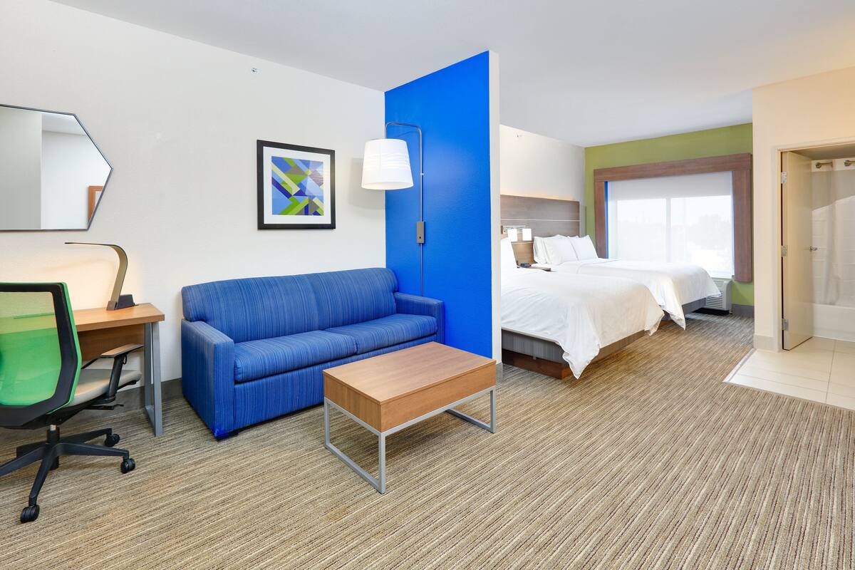 A hotel room with a blue accent wall, a bed, a desk with a chair, a coffee table, and a large window.
