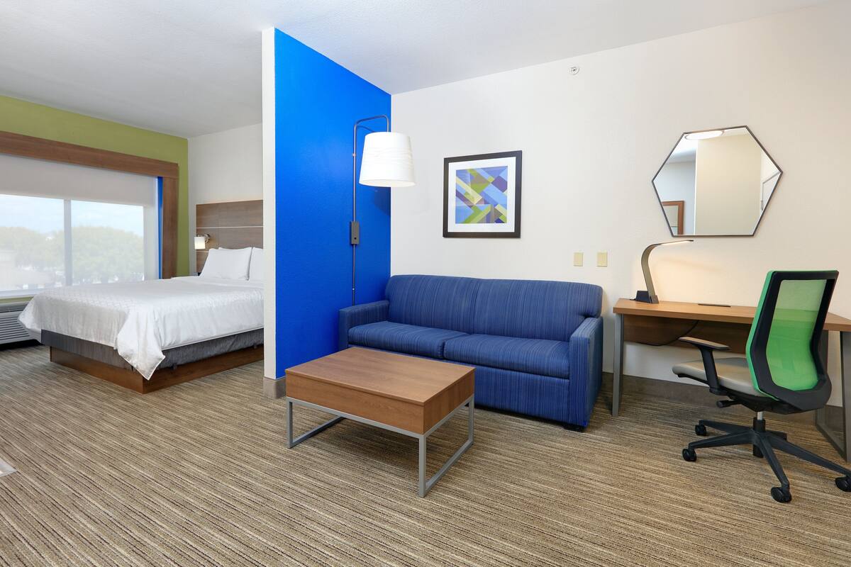 A hotel room with a bed, a blue sofa, a wooden coffee table, a desk with a chair, a lamp, and a mirror.