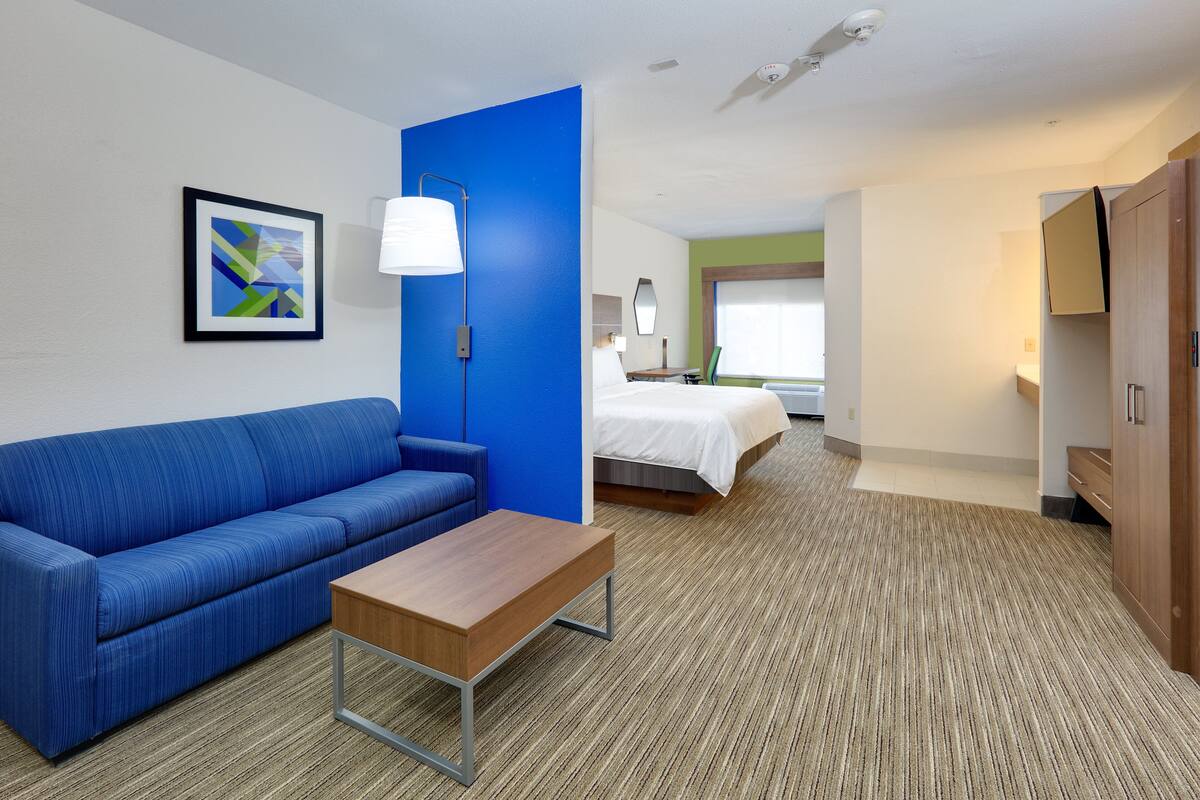 A hotel room with a blue sofa, a wooden table, a bed with white bedding, a blue accent wall, a lamp, and a framed picture on the wall.