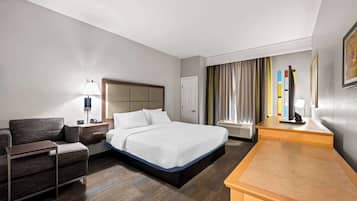 Une chambre d’hôtel avec un lit, un bureau, une chaise, une table de chevet, une lampe, une fenêtre avec des rideaux et un tableau encadré au mur.