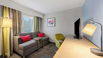 A modern hotel room with a sofa, a green chair, a flat-screen TV, and a desk with a lamp.