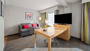 Une chambre d’hôtel avec un bureau en bois, un canapé gris avec des coussins rouges, une télévision et une grande fenêtre avec des rideaux.