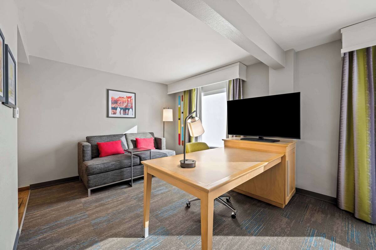 A hotel room with a wooden desk, a grey sofa with red cushions, a television, and a large window with curtains.