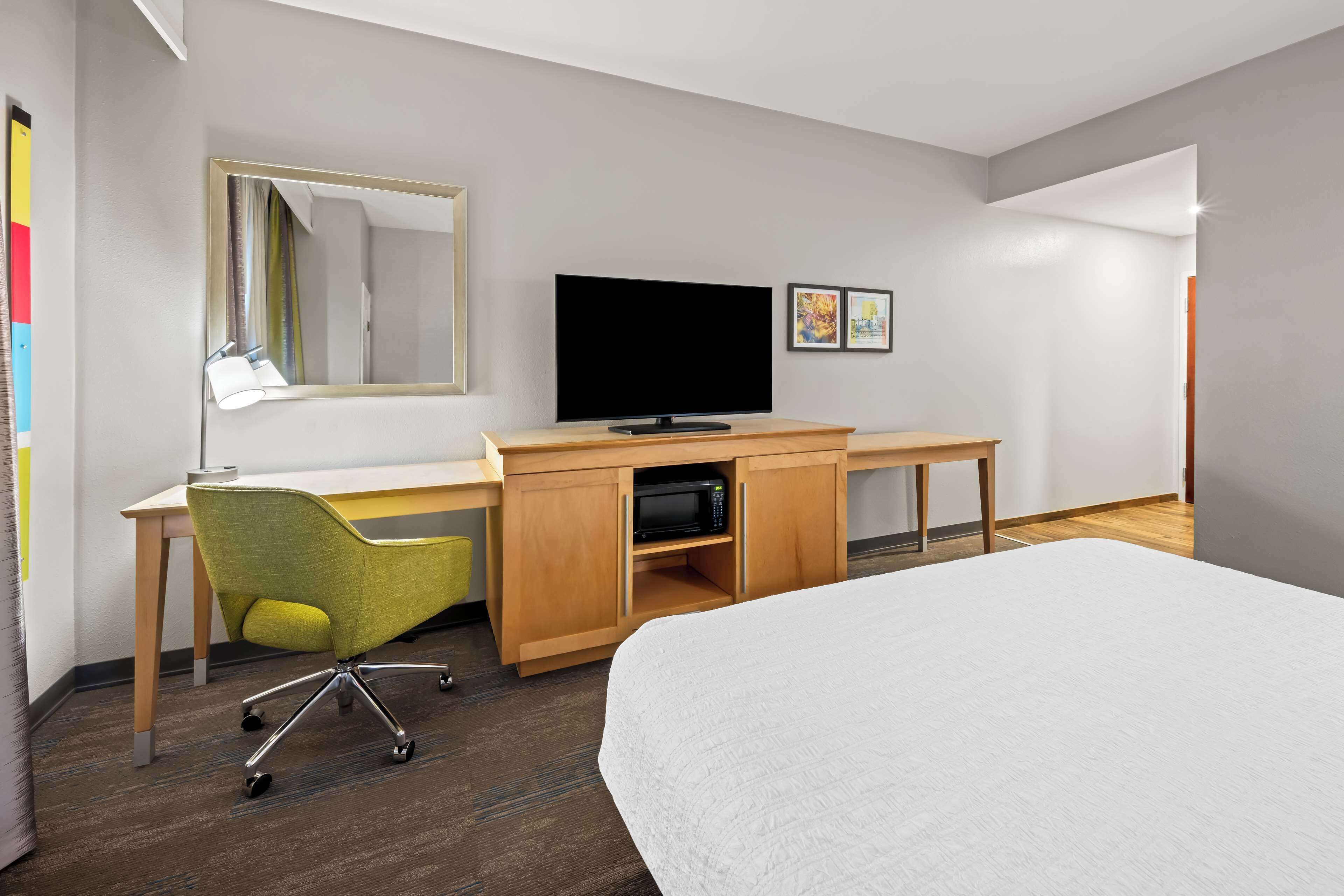 A hotel room with a bed, a desk with a computer and a TV, a chair, and a mirror.