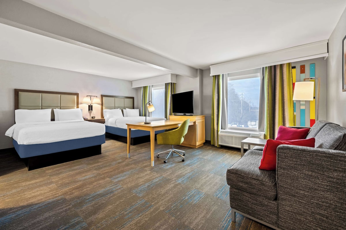 A hotel room with two beds, a desk, a chair, a sofa, and a TV.