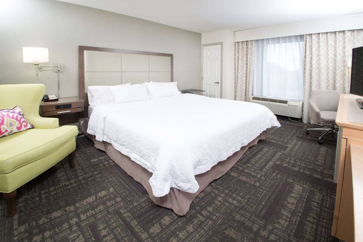 A hotel room with a large bed, a desk, a chair, a lamp, and a window with curtains.