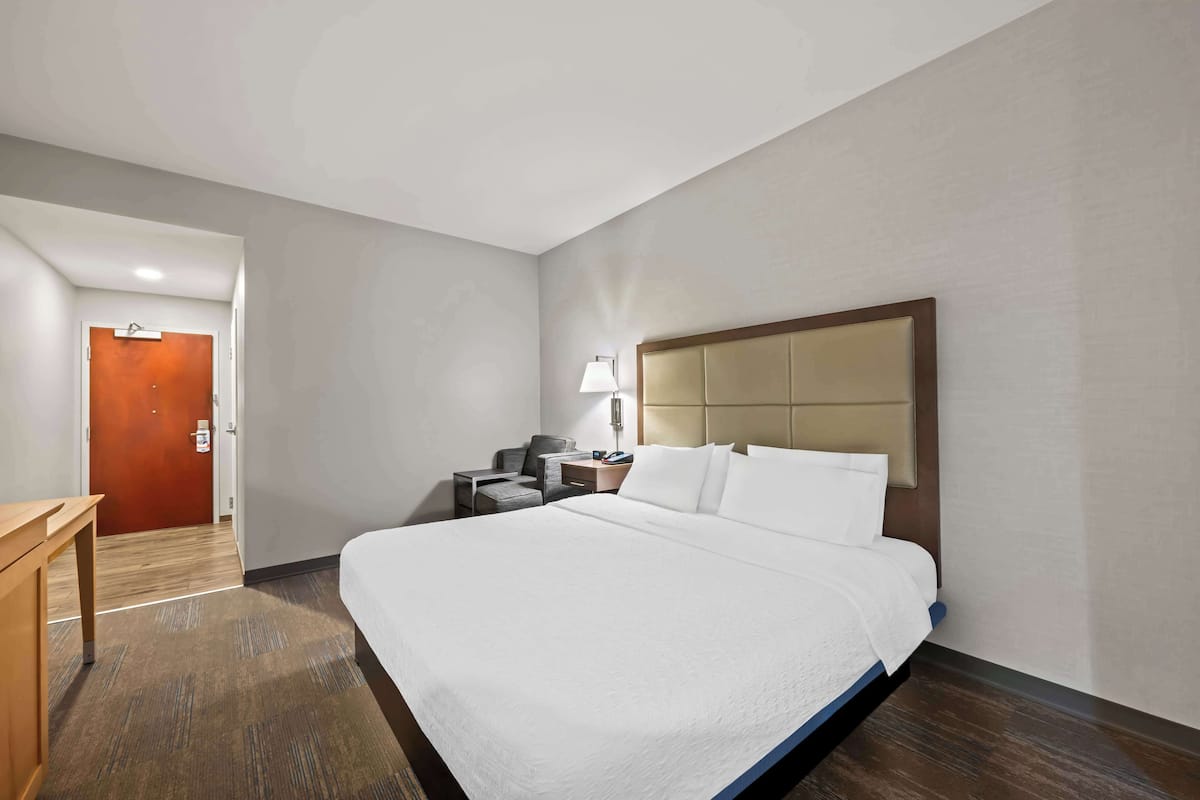 A hotel room with a large bed, a desk, a chair, a lamp, and a door.