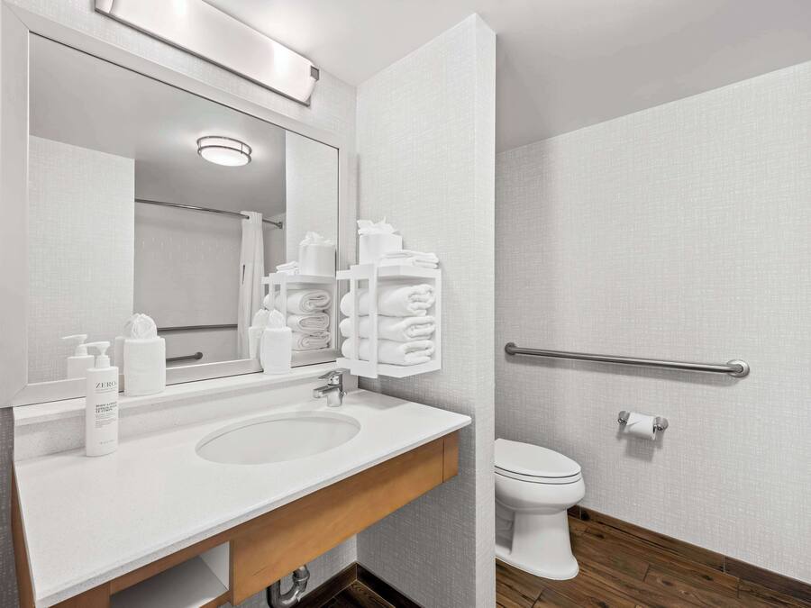 A modern bathroom with a white sink, mirror, and toilet. There are toiletries on the sink and a towel rack above the toilet.