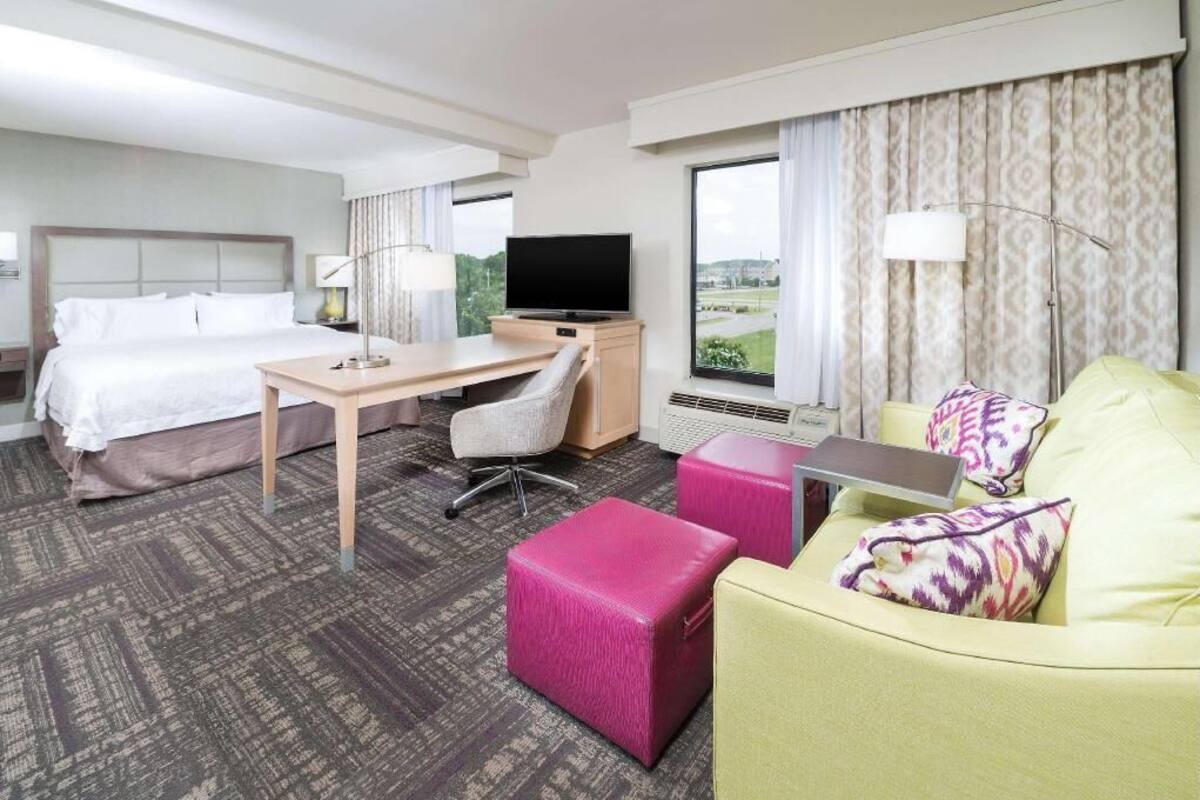 A hotel room with a bed, desk, chair, ottoman, sofa, TV, and large windows.