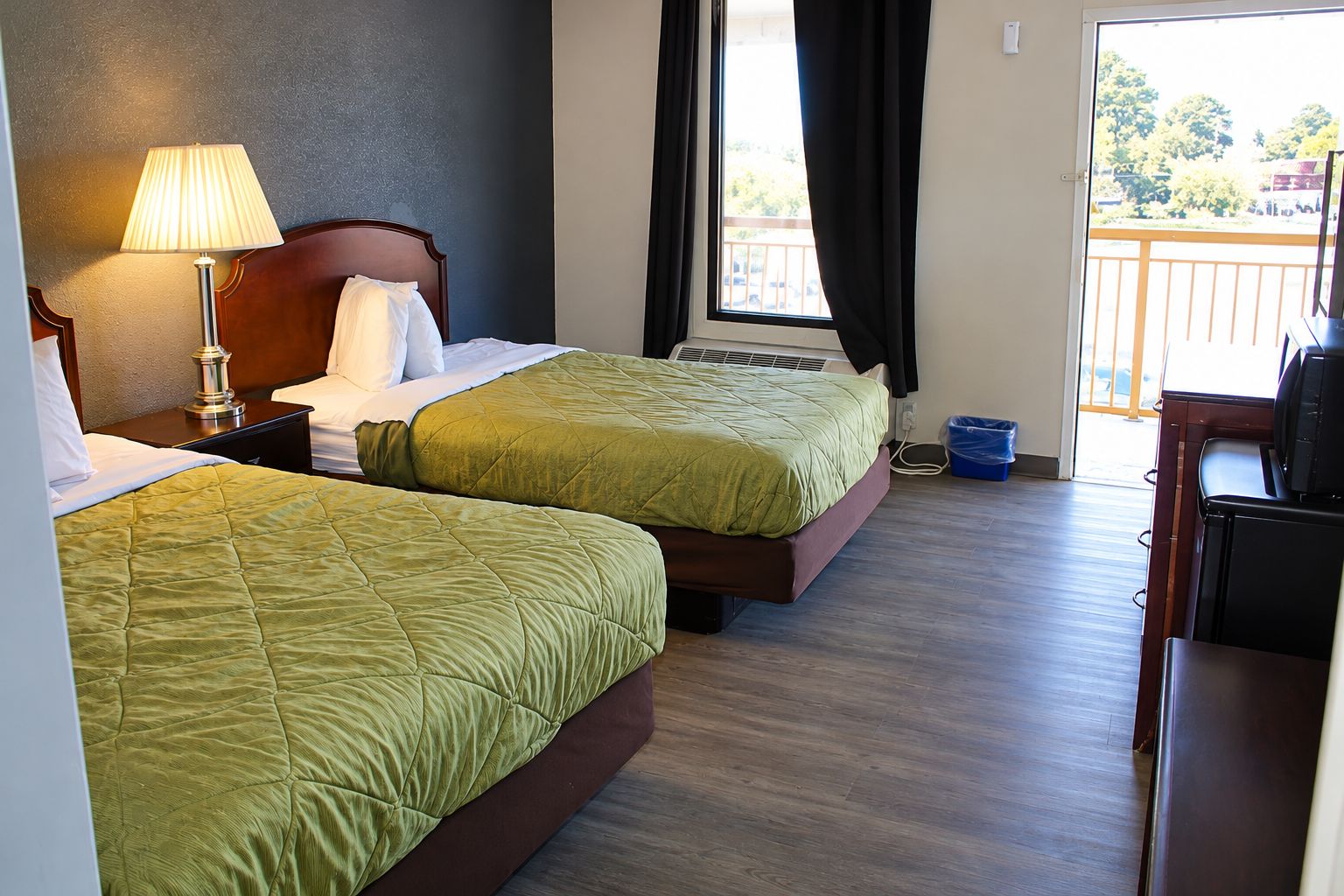 A hotel room with two beds, a desk, a lamp, and a view of the outdoors.