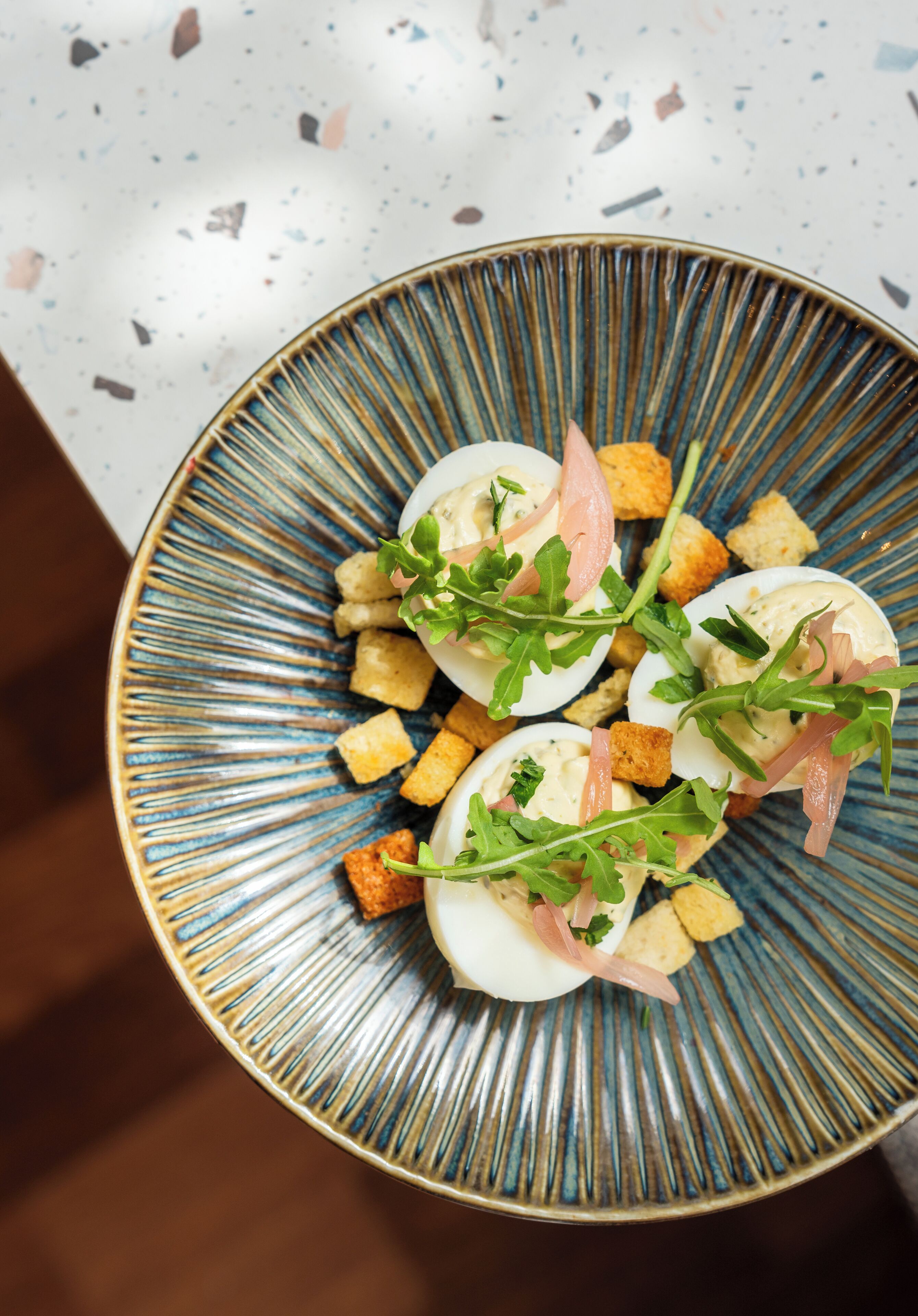 Une assiette d’œufs mimosa décorée de roquette et de lardons, présentée sur une assiette aux motifs bleus et dorés.