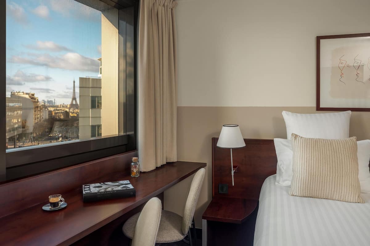 A hotel room with a bed, a desk, a chair, a nightstand, a lamp, and a window with a view of buildings.