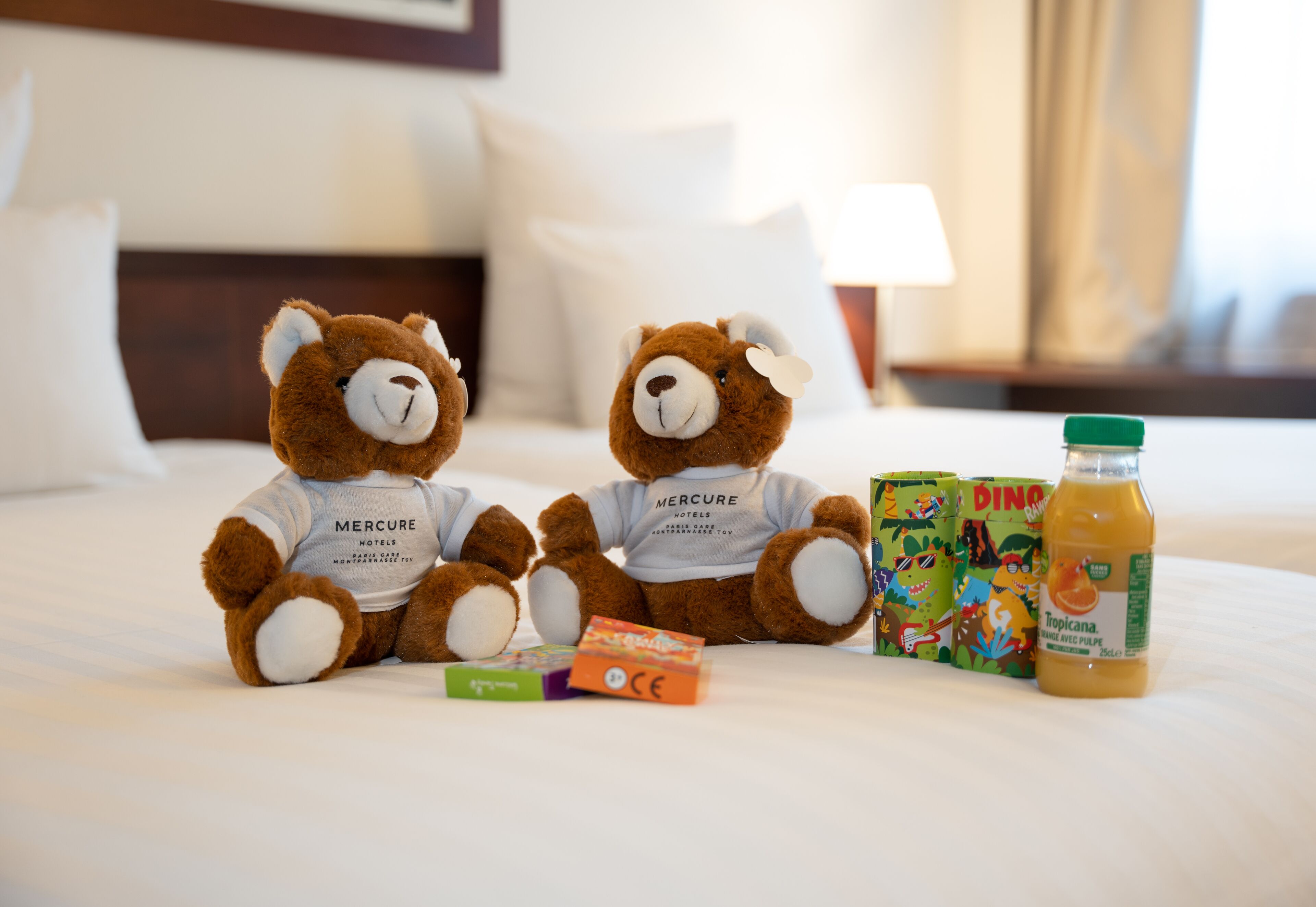 Two plush bears on a bed with a bottle of Tropicana juice, a toy box, and a small orange box.