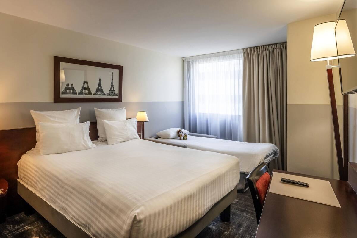 A hotel room with two beds, a desk, a lamp, a framed picture of the Eiffel Tower, and a window with curtains.